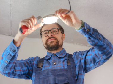 Elektrikçi iş başında. Elektrik kablolarının tamiri ve tavan lambalarının değiştirilmesi için servis. İnşaatçı tavana küçük bir lamba yerleştiriyor. Kiralık centilmen ev işlerine yardım eder.