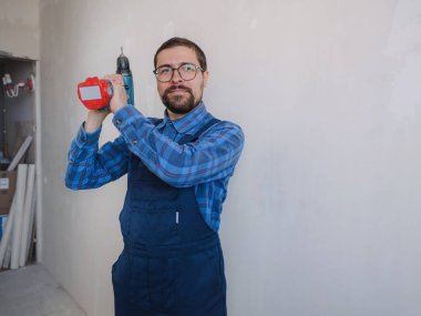 Mavi takım elbiseli genç adam apartman dairesinde çalışıyor. Ev yenileme konsepti. Elinde elektrikli matkap tutan tamirci beyaz duvar arkasında izole edilmiş. Enstrümanlar aksesuarlar yenileme dairesi