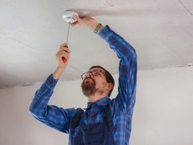 Mavi takım elbiseli genç elektrikçi tavana duman dedektörü yerleştiriyor.