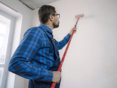 Mavi takım elbiseli genç adam beyaz duvar odası arka planında boya silindiri tutuyor. Yenileme dairesi için aksesuarlar. Ev konseptini onar