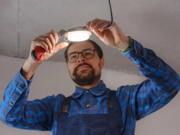 Elektrikçi iş başında. Elektrik kablolarının tamiri ve tavan lambalarının değiştirilmesi için servis. İnşaatçı tavana küçük bir lamba yerleştiriyor. Kiralık centilmen ev işlerine yardım eder.