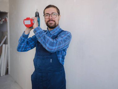 Mavi takım elbiseli genç adam apartman dairesinde çalışıyor. Ev yenileme konsepti. Elinde elektrikli matkap tutan tamirci beyaz duvar arkasında izole edilmiş. Enstrümanlar aksesuarlar yenileme dairesi