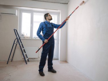 Mavi takım elbiseli genç adam beyaz duvar odası arka planında boya silindiri tutuyor. Yenileme dairesi için aksesuarlar. Ev konseptini onar