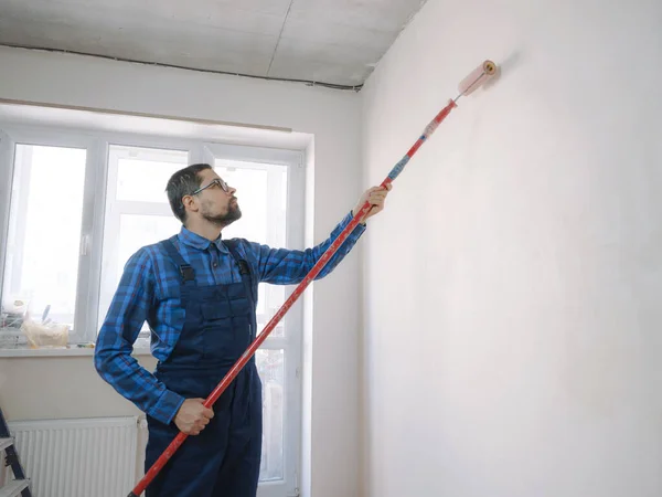 young man in blue work suit hold paint roller, over white wall room background. Instruments accessories for renovation apartment room. Repair home concept