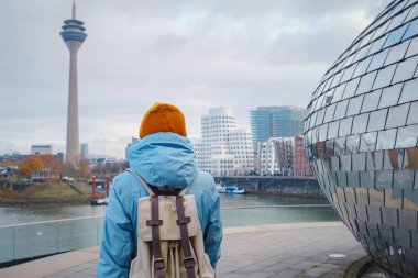 autumn or winter travel to Dusseldorf, Germany. young Asian tourist or student in blue jacket and yellow hat  symbol of Ukraine walks through sights of European city. beautiful view in the Media Bay