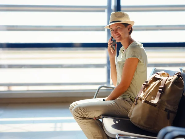 young asian female traveler with hipster backpack and hat at airport waiting for departure at Vienna Airport. Traveling woman Remote Work Online on Mobile Phone in Boarding Lounge of Airline Hub