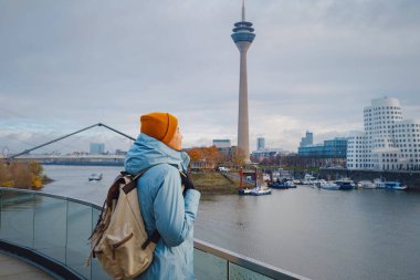 autumn or winter travel to Dusseldorf, Germany. young Asian tourist or student in blue jacket and yellow hat  symbol of Ukraine walks through sights of European city. beautiful view in the Media Bay