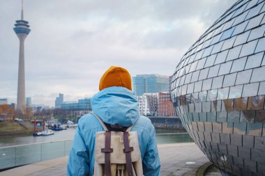 autumn or winter travel to Dusseldorf, Germany. young Asian tourist or student in blue jacket and yellow hat  symbol of Ukraine walks through sights of European city. beautiful view in the Media Bay