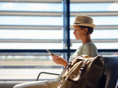 young asian female traveler with hipster backpack and hat at airport waiting for departure at Vienna Airport. Traveling woman Remote Work Online on Mobile Phone in Boarding Lounge of Airline Hub