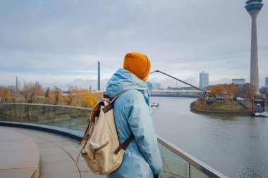 autumn or winter travel to Dusseldorf, Germany. young Asian tourist or student in blue jacket and yellow hat  symbol of Ukraine walks through sights of European city. beautiful view in the Media Bay
