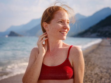 Tropikal plajda kırmızı bikinili güzel kızıl kadın. Denizde gülümseyen genç bayanın portresi. Kızıl saçlı, çilli, güneşin tadını çıkaran mutlu bir kız. Güneş ile güvenli etkileşim kavramı