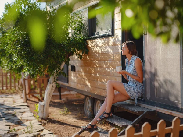 Woman is drinking coffee , morning routine near her Tiny house. First property. Small apartment in summer garden. Minimalism. Moving in. Living alone. Charming trailer house