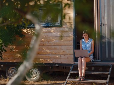 Dizüstü bilgisayarı olan mutlu genç Asyalı kadın dışarıda, küçük bir evin yanında dinleniyor, hafta sonu uzakta ve ücra ofis fikri. Küçük evler ve Küçük Yaşayanlar konsepti. Yaz bahçesi