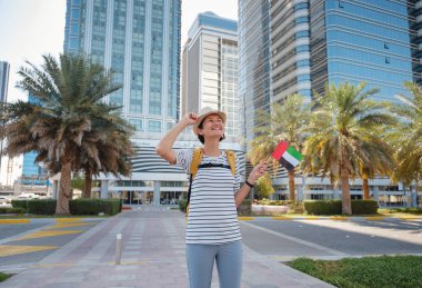 Abu Dabi 'deki manzaralı gökdelenlere karşı sırt çantalı ve şapkalı mutlu Asyalı genç bayan gezgin. Göçmenlik, öğrenci eğitimi ve Arap Emirlikleri vatandaşlık kavramı