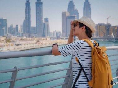 Şapkalı ve sarı sırt çantalı Asyalı mutlu gezgin Dubai Creek Kanalı 'nın büyüleyici panoramik manzarasının ve Tolerance Köprüsü' nün yanından geçen ünlü gökdelen Burj Khalifa 'nın keyfini çıkarıyor.