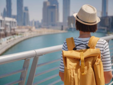 Şapkalı ve sarı sırt çantalı Asyalı mutlu gezgin Dubai Creek Kanalı 'nın büyüleyici panoramik manzarasının ve Tolerance Köprüsü' nün yanından geçen ünlü gökdelen Burj Khalifa 'nın keyfini çıkarıyor.