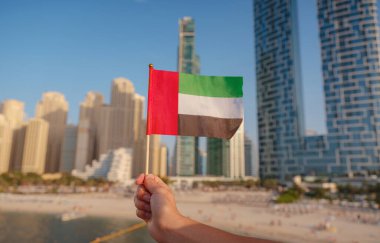 Close up of a female hand holding tiny flag of UAE against Dubai skyline. Photo of UAE national day celebration spirit. clipart