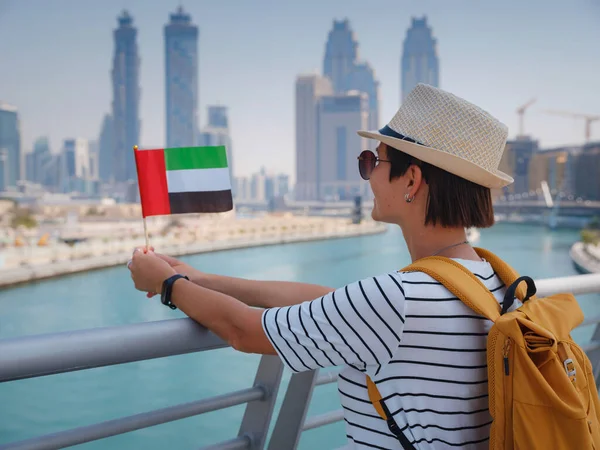 アラブ首長国連邦の旗を持つ帽子と黄色のバックパックを持つアジアの幸せな旅行者の女性は ドバイクリーク運河と有名な高層ビルブルジュ ハリファの素晴らしいパノラマビューを楽しんでいます — ストック写真