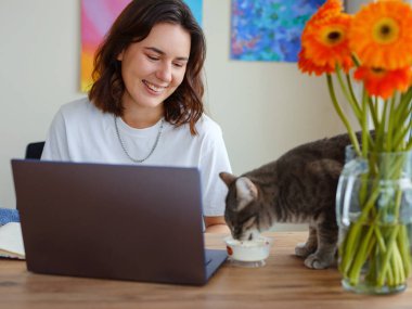 Genç kadın dizüstü bilgisayarda evden çalışıyor. Oturma odasındaki evcil hayvanla internetten çalış ya da çalış. Masada kedi yiyor..