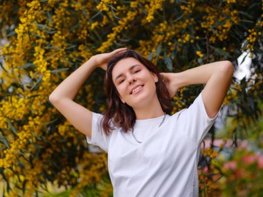 Koyu kıvırcık saçlı, mükemmel tenli beyaz tişörtlü çiçek açan mimoza çiçeklerinin yanında poz veren güzel bir genç kadın. Çıplak makyaj malzemesi. Alerji sezonu, antihistaminler. Aroma doğal parfümleri yağlar.