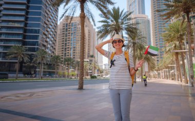 Birleşik Arap Emirlikleri 'nde iyi yolculuklar. Üzerinde Birleşik Arap Emirlikleri bayrağı olan sarı sırt çantalı genç bir kadın güneşli yaz gününde Dubai 'de yürüyor..