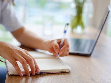 Laptop kullanan ve evde çalışırken gülümseyen gündelik giysili güzel Latin bir kadın. Öğrencilerin dizüstü bilgisayar masasında yönetici olmaları için eğitim notları yazıyorlar.