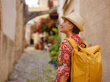 Kırmızı elbiseli ve sırt çantalı genç Asyalı kadın eski şehrin rahat dar sokaklarına bakıyor. Turizm, tatil ve keşif konsepti, Güney Avrupa 'yı ziyaret eden kadın gezgin, Rodos adası Yunanistan