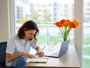 Laptop kullanan ve evde çalışırken gülümseyen gündelik giysili güzel Latin bir kadın. Öğrencilerin dizüstü bilgisayar masasında yönetici olmaları için eğitildikleri notları yazıyorlar.