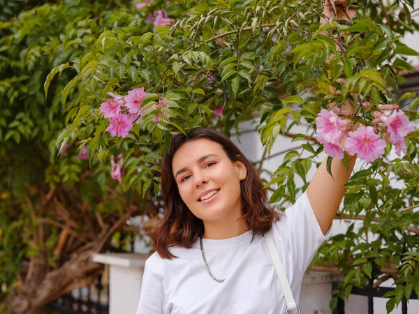 stock image Beautiful young woman with dark curly hair and perfect skin wearing white tshirt posing near blooming flowers in garden. Nude make up. Allergy season, antihistamines. Aroma oils natural perfumes