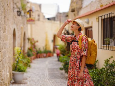 Kırmızı elbiseli ve sırt çantalı genç Asyalı kadın eski şehrin rahat dar sokaklarına bakıyor. Turizm, tatil ve keşif konsepti, Güney Avrupa 'yı ziyaret eden kadın gezgin, Rodos adası Yunanistan