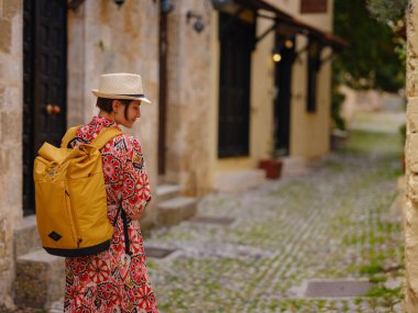 Kırmızı elbiseli ve sırt çantalı genç Asyalı kadın eski şehrin rahat dar sokaklarına bakıyor. Turizm, tatil ve keşif konsepti, Güney Avrupa 'yı ziyaret eden kadın gezgin, Rodos adası Yunanistan