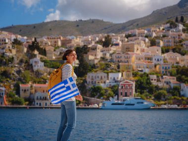 Sırt çantalı mutlu Asyalı kadın ve Yunan bayrağı Symi Adaları 'nda tatilinin tadını çıkarıyor. Liman manzarası, Yunanistan 'ın Dodecanese kentindeki küçük bir ada, sakin bir atmosfer ve muhteşem bir mimari..