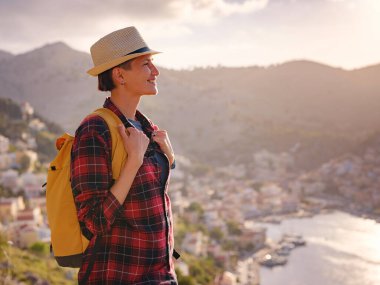 Sırt çantalı mutlu Asyalı kadın gün batımında Symi Adaları 'nda tatilin tadını çıkarıyor. Liman manzarası, Yunanistan 'ın Dodecanese kentindeki küçük bir ada, sakin bir atmosfer ve muhteşem bir mimari..