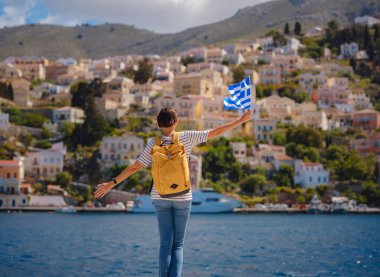 Sırt çantalı mutlu Asyalı kadın ve Yunan bayrağı Symi Adaları 'nda tatilinin tadını çıkarıyor. Liman manzarası, Yunanistan 'ın Dodecanese kentindeki küçük bir ada, sakin bir atmosfer ve muhteşem bir mimari..