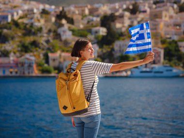 Sırt çantalı mutlu Asyalı kadın ve Yunan bayrağı Symi Adaları 'nda tatilinin tadını çıkarıyor. Liman manzarası, Yunanistan 'ın Dodecanese kentindeki küçük bir ada, sakin bir atmosfer ve muhteşem bir mimari..