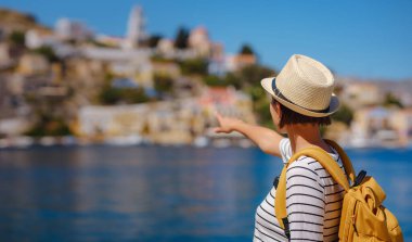 Sırt çantalı mutlu Asyalı kadın Symi Adaları 'nda tatilinin tadını çıkarıyor. Liman manzarası, Yunanistan 'ın Dodecanese kentindeki küçük bir ada, sakin bir atmosfer ve muhteşem bir mimari..