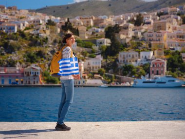 Sırt çantalı mutlu Asyalı kadın ve Yunan bayrağı Symi Adaları 'nda tatilinin tadını çıkarıyor. Liman manzarası, Yunanistan 'ın Dodecanese kentindeki küçük bir ada, sakin bir atmosfer ve muhteşem bir mimari..