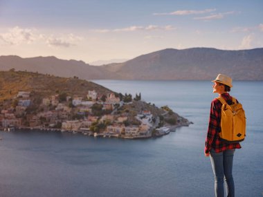 Sırt çantalı mutlu Asyalı kadın gün batımında Symi Adaları 'nda tatilin tadını çıkarıyor. Liman manzarası, Yunanistan 'ın Dodecanese kentindeki küçük bir ada, sakin bir atmosfer ve muhteşem bir mimari..