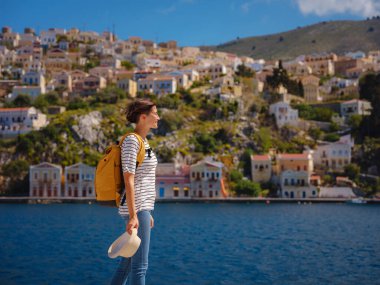 Sırt çantalı mutlu Asyalı kadın Symi Adaları 'nda tatilinin tadını çıkarıyor. Liman manzarası, Yunanistan 'ın Dodecanese kentindeki küçük bir ada, sakin bir atmosfer ve muhteşem bir mimari..