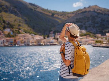 Sırt çantalı mutlu Asyalı kadın Symi Adaları 'nda tatilinin tadını çıkarıyor. Liman manzarası, Yunanistan 'ın Dodecanese kentindeki küçük bir ada, sakin bir atmosfer ve muhteşem bir mimari..
