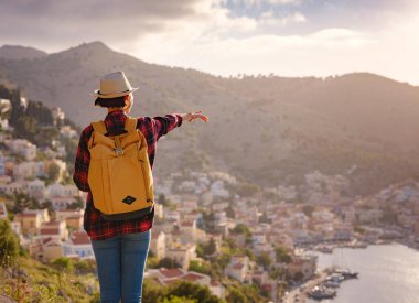 Sırt çantalı mutlu Asyalı kadın gün batımında Symi Adaları 'nda tatilin tadını çıkarıyor. Liman manzarası, Yunanistan 'ın Dodecanese kentindeki küçük bir ada, sakin bir atmosfer ve muhteşem bir mimari..