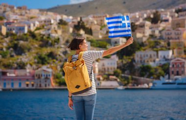 Sırt çantalı mutlu Asyalı kadın ve Yunan bayrağı Symi Adaları 'nda tatilinin tadını çıkarıyor. Liman manzarası, Yunanistan 'ın Dodecanese kentindeki küçük bir ada, sakin bir atmosfer ve muhteşem bir mimari..