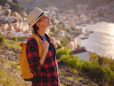Sırt çantalı mutlu Asyalı kadın gün batımında Symi Adaları 'nda tatilin tadını çıkarıyor. Liman manzarası, Yunanistan 'ın Dodecanese kentindeki küçük bir ada, sakin bir atmosfer ve muhteşem bir mimari..