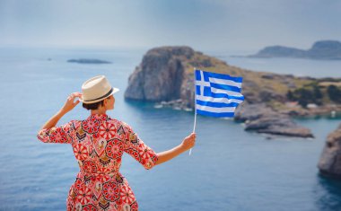Yunan Adaları 'nda güneşli bir günün tadını çıkaran güzel mutlu kadın. Yunanistan 'a seyahat, turizm sezonu dışında Akdeniz adaları. Ulusal Yunan bayrağıyla gezen genç kadın deniz manzarasının keyfini çıkarıyor