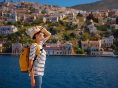 Sırt çantalı mutlu Asyalı kadın Symi Adaları 'nda tatilinin tadını çıkarıyor. Liman manzarası, Yunanistan 'ın Dodecanese kentindeki küçük bir ada, sakin bir atmosfer ve muhteşem bir mimari..