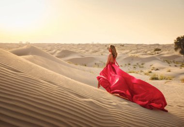 Çöl macerası. Gün batımında BAE çölünün kumullarında kırmızı ipek elbiseli genç bir Arap kadın, muhteşem bir manzara. Dubai Çölü Koruma Alanı, Birleşik Arap Emirlikleri.