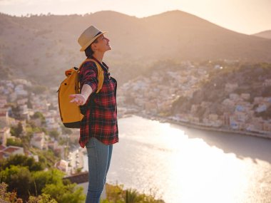 Sırt çantalı mutlu Asyalı kadın gün batımında Symi Adaları 'nda tatilin tadını çıkarıyor. Liman manzarası, Yunanistan 'ın Dodecanese kentindeki küçük bir ada, sakin bir atmosfer ve muhteşem bir mimari..