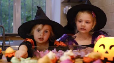 Cadılar Bayramı için mutfakta kostüm giymiş iki kız Jack-o-Lantern 'le oturuyor, gülüyor, tatlı ve kurabiye tadıyor. Mutlu çocuklar Cadılar Bayramı için hazırlanıyor. Cadılar Bayramı için hazırlanılıyor.