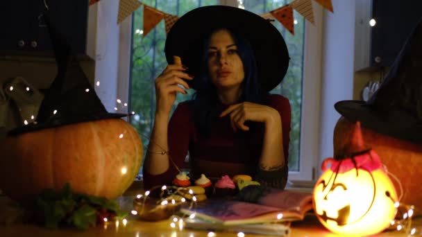 可怕的美丽女人女巫在厨房里庆祝万圣节 南瓜和花环在厨房里 戴女巫帽的女人在准备万圣节派对 女士品尝饼干和纸杯蛋糕 — 图库视频影像