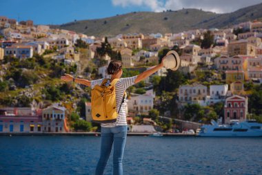 Sırt çantalı mutlu Asyalı kadın Symi Adaları 'nda tatilinin tadını çıkarıyor. Liman manzarası, Yunanistan 'ın Dodecanese kentindeki küçük bir ada, sakin bir atmosfer ve muhteşem bir mimari..
